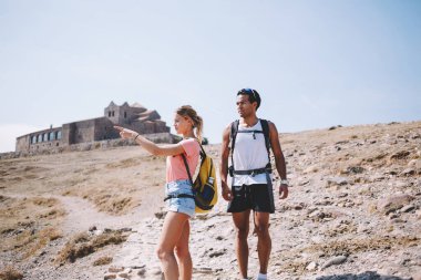 Güneşli yaz gününde kayalık zeminde birlikte yürüyen genç adam ve kadın sırt çantalarıyla seyahat ederken ve arka planda binayla birlikte günlük giysiler giyerken.
