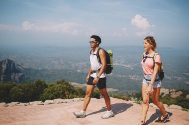 Güneşli bir günde sırt çantaları ve yaz kıyafetleri giyerek tepede birlikte yürüyen çok ırklı genç bir çift.