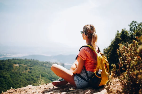 穿着夏装 戴着太阳镜 背着背包坐在山顶上欣赏风景如画的年轻女性游客的全景背影 — 图库照片