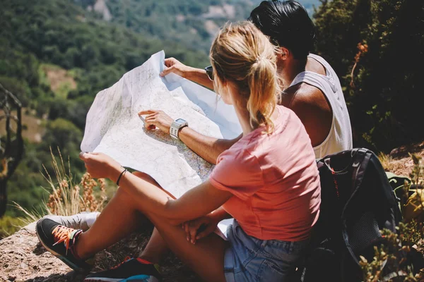 年轻的无名游客坐在山顶上 戴着太阳镜 身穿休闲装 在阳光普照的天气里 一边探索地图 一边回头看 — 图库照片