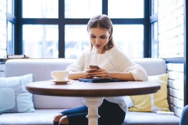 Odaklanmış, uzun saçlı, süveter giyen, telefonla konuşan ve gündüz vakti açık kafede otururken sıcak içecekle masaya oturan bir kadın.