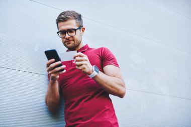 Gri duvara yaslanmış günlük giysiler ve gözlük takan ve akıllı telefondan telefon numarasını çeviren konsantre sakallı adamdan.