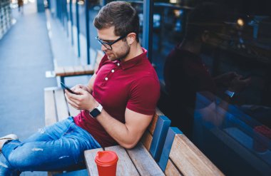 Düşünceli, sakallı, Polo tişörtlü, tahta bankta kırmızı fincan kahveyle oturan ve şehir planında akıllı telefon sörfü yapan adamdan.