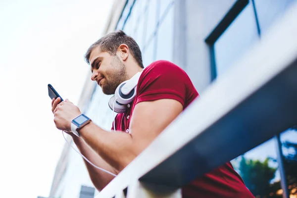 Positive Young Unshaven Guy Smart Watch Headphones Neck Browsing Smartphone — Stock Photo, Image
