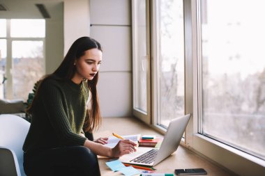 Kendine güveni tam bir kadın. Pencere kenarında oturmuş elinde ağ defteri ve ofis malzemeleriyle ders çalışırken elinde kalem tutarken ekrana bakıyor.