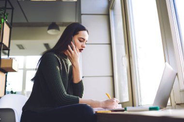 Pencerenin kenarında dizüstü bilgisayarla oturan ve iş yerindeki cep telefonuyla konuşurken notlar yazan kendini ifade eden kadın girişimcinin düşük açısı