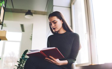 Dışa dönük kadın öğrencilerden biri pencerenin kenarında oturmuş evde dinlenirken kırmızı kapaklı ilginç bir kitap okuyor.
