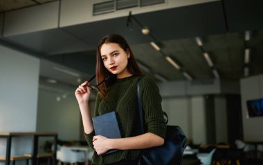 Yeşil kazaklı ve siyah gözlüklü bir bayan öğrenci modern kantinde bulanık arka planda kameraya bakıyor.