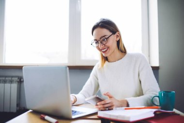 Neşeli genç bayan yönetici sosyal medyada arkadaşıyla sohbet ediyor kahve molasında dizüstü bilgisayarını kullanıyor bardak ve defterle masada oturuyor.