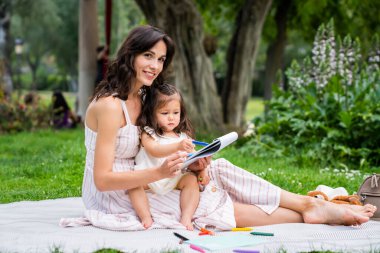 Neşeli, kıvırcık, siyah saçlı bir kadın çimenlerin üzerinde bacak bacak üstüne atarak oturuyor ve küçük bir kız pastel kalemle kalemle eğleniyor ve dışarıdaki kameraya bakıyor. 