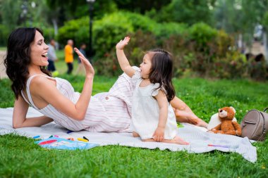 Koyu saçlı, neşeli bir kadın parkta battaniyede dinleniyor ve beyaz elbiseli güzel kıza beşlik çakıyor.