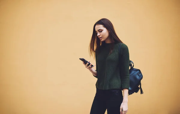 携帯電話を見ている間に手やテキストメッセージでスマートフォンに立ってバックパック付きのカジュアルなセーターやジーンズを身に着けている若い女性 — ストック写真
