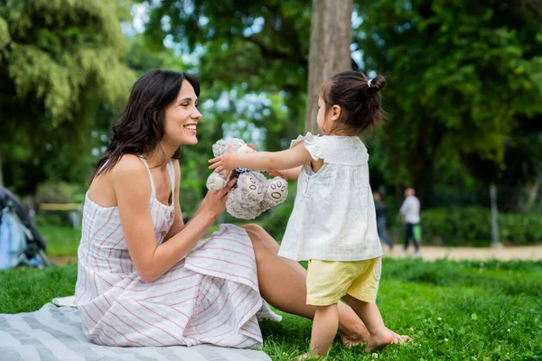 Gülümseyen annenin battaniyeye oturup, parkta piknikte çimenlerde yürüyen sevimli çocuğa yumuşak gri oyuncak ayıyı önermesi. 