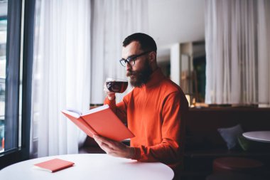 Konsantre olmuş erkek, açık modern kafede tek başına vakit geçirirken elinde çay fincanı ile oturmuş kitap okuyor.