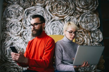 Yoğunlaşmış meslektaşlar modern dizüstü bilgisayara göz atıyorlar ve proje için akıllı telefon bulma bilgilerini dekore edilmiş duvarlarla aydınlatma odasında vakit geçirirken kullanıyorlar.