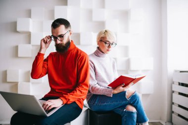 Gözlük takan, dizüstü bilgisayara dokunan serbest çalışan bir erkek ve açık not defteriyle oturan ciddi bir kadın birlikte modern bir iş yerinde çalışıyorlar.