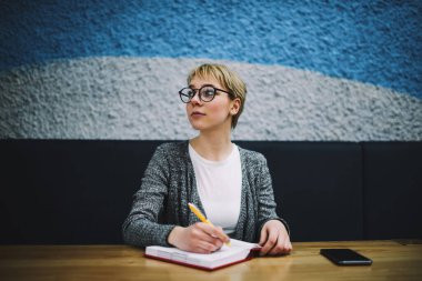 Gündelik giysiler içinde ciddi bir kadın serbest çalışan. Çalışırken cep telefonu notları yazarak masaya bakıyor.