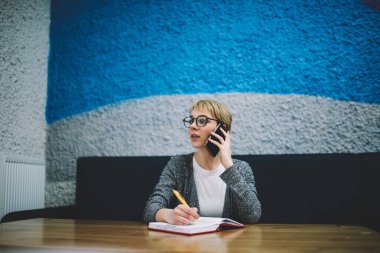 Düşük açılı, kısa saçlı ve gözlüklü bir kadın elinde not defteriyle masada oturmuş akıllı telefonuyla sohbet ediyor.