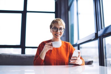 Düşük açılı, dişlek gülümsemeli, pencerenin yanındaki masaya bakan kahve içen ve akıllı telefondan arkadaşlarıyla sohbet eden mutlu bir kadın.