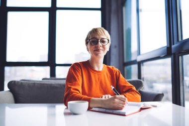 Gülümseyen uzaktan kumandalı kadın işçilerin gündelik kıyafetleriyle masaya oturup kahve içerek tek başlarına çalışırken notlar almalarından.