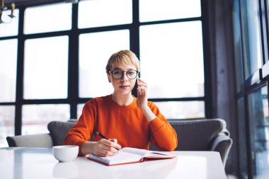 Genç, gülümseyen kadın, cep telefonuyla konuşuyor. Kafeteryada defter ve bir fincan sıcak içecekle masada oturuyor.