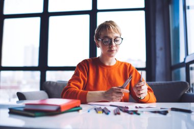 Düşük açılı, günlük kıyafetler giyen, gözlüklü, not defterli, iş sırasında renkli kalem seçen kadın sanatçı.