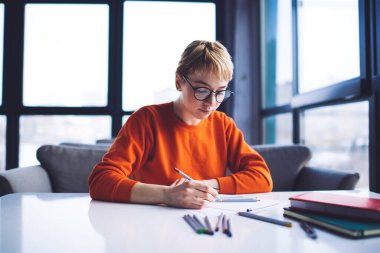 Gözlüklü, kısa saçlı, kurşun kalemle masaya oturan ve iş sırasında çizim yapan serbest çalışan genç bir kadın.