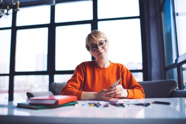 Gündelik kıyafetlerle gülümseyen kadın, dinlenirken pencerenin yanındaki masada renkli kalemlerle kameraya bakıyor.
