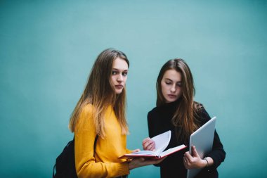 Düşük açılı düşünceli bayan öğrenciler dizüstü bilgisayarlarıyla turkuaz duvarın yanında duruyor ve yeni ev projesini tartışırken defteri açıyorlar.