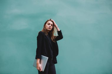 Düşük açılı, sıradan giyinmiş, turkuaz arka planda modern dizüstü bilgisayarla saçlarına dokunan kameraya bakan düşünceli bir bayan öğrenci.