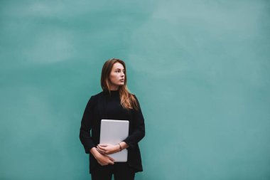 Uzun saçlı, şık siyah kıyafetli, modern bilgisayarla turkuaz duvarın yanında duran düşünceli bir bayan öğrenci.