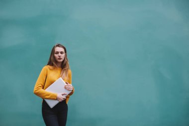 Uzun saçlı, düşünceli bir bayan öğrenci elinde dizüstü bilgisayarla turkuaz duvarın yanında boş vakti varken kameraya bakıyor.