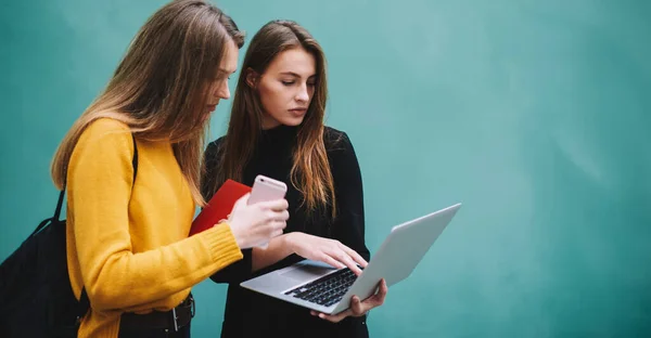 许多严肃的女学生站在绿松石墙边 一边拿着手机一边用笔记本电脑 一边一起学习 — 图库照片
