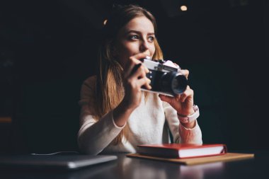 Sıradan giyinen karizmatik kadın fotoğraf makinesine fotoğraf çektirip siyah odada vakit geçirirken pencereden dışarı bakıyor.