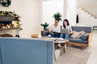 Oturma odasındaki minderlerle kanepede vakit geçirirken projede birlikte çalışarak dizüstü bilgisayara bakan gülümseyen kadınlar.