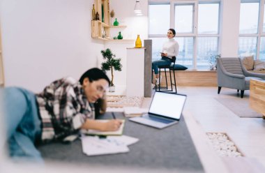 Keyifli bir kadın, gündelik giysiler içinde masada otururken arkadaşı da notlar yazıyor ve modern çalışma sahasında rahat bir iç mekan ile dizüstü bilgisayar kullanıyor.