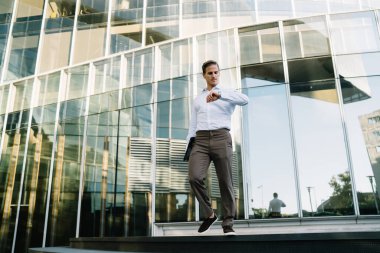 Beyaz iş adamı 30 yaşında, ofis yakınlarındaki zamanı kontrol ediyor. Finansal bölgedeki iş molasında modern teknolojiyi kullanıyor. Giyilebilir ekipmanlara bakan zeki, sıradan bir erkek girişimci.