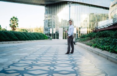 Modern mobil cihazlı akıllı girişimcinin tam boy portresi finans bölgesinde şehir merkezinde duruyor ve kameraya bakıyor, beyaz iş adamı şehir merkezinde poz veriyor.