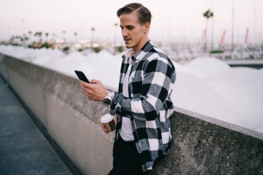 Yakışıklı beyaz tenli hippi, açık havada akıllı telefon haberleri için internet sayfası kullanıyor, 20 'li yaşlardaki genç hippi blogcu elinde kahve fincanı ile cep telefonuyla posta gönderiyor.
