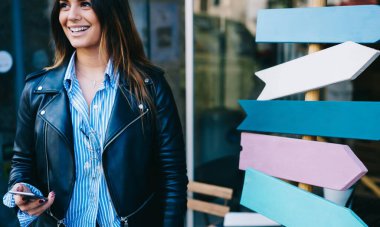 Dişli tebessümlü genç bir kadın renkli tabelaların yanında dikilip caddenin bulanık arka planında cep telefonuna bakıyor.