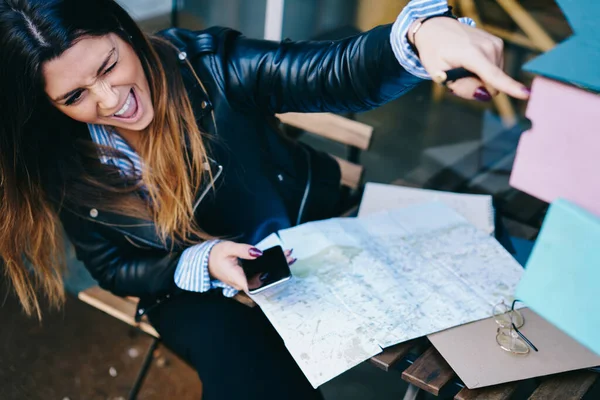 Happy Woman Sitting Table Map Mobile Phone Pointing Away Winking — Stockfoto