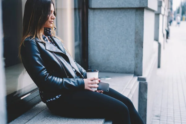 在街上的水泥窗台上 坐着时尚女性的侧面景观 她带着纸杯热饮和手机 — 图库照片