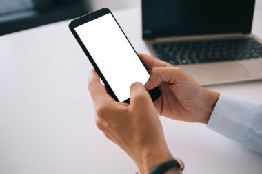 Gömlekli yüzü olmayan, boş ekranlı akıllı telefonu karıştıran ve ofiste proje üzerinde çalışırken dizüstü bilgisayarlı bir masada oturan kişi.