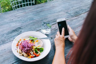 Anonim kadın blogcu akıllı telefon kullanıyor ve gün içinde kafede lezzetli salata ve bir bardak suyla ahşap masada oturuyor.