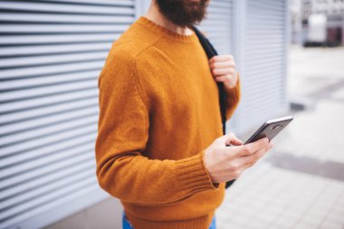 Sırt çantalı sakallı, sıradan bir erkek duvarın yanında duruyor. Şehirde sokakta yürürken modern cep telefonlarına bakıyor.
