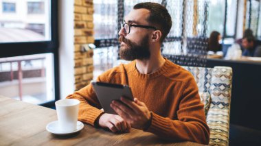 Ciddi sakallı bir adam, günlük süveter ve gözlüklerle kafede oturmuş, elinde bir bardak sıcak içecekle, kafede tablet üzerinde çalışırken pencereden dışarı bakıyor.