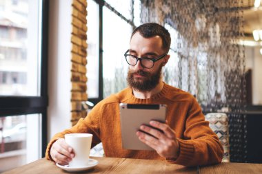 Konsantre sakallı erkek serbest çalışan pencerenin yanındaki masada elinde bir kupa sıcak içecekle rahat bir kafede çalışırken tablet kullanıyor.