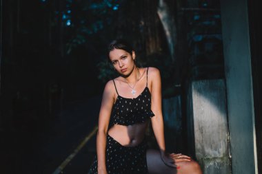 Sakin İspanyol kadın, siyah yazlık bluz ve etek giymiş. Alacakaranlıkta ormanda tuğla binanın önünde dikilirken kameraya bakıyor.