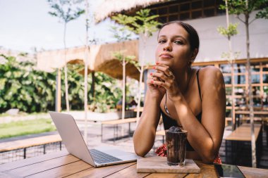 Gündüz vakti modern açık hava kafesinde içki ve dizüstü bilgisayarla masada oturan, dirseklerine yaslanan ve çene dokunan genç bir kadın hayal kurarak kameraya bakıyor.
