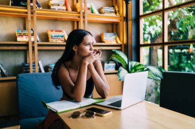 Çekici genç kız öğrenci netbook ile masada oturuyor ve hoş düşüncelerle bakışlarını kaçırarak çalışma alanında defterini açıyor.